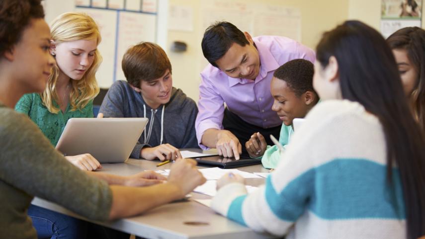 A teacher assisting a group of diverse high school students working together on a project at a table.